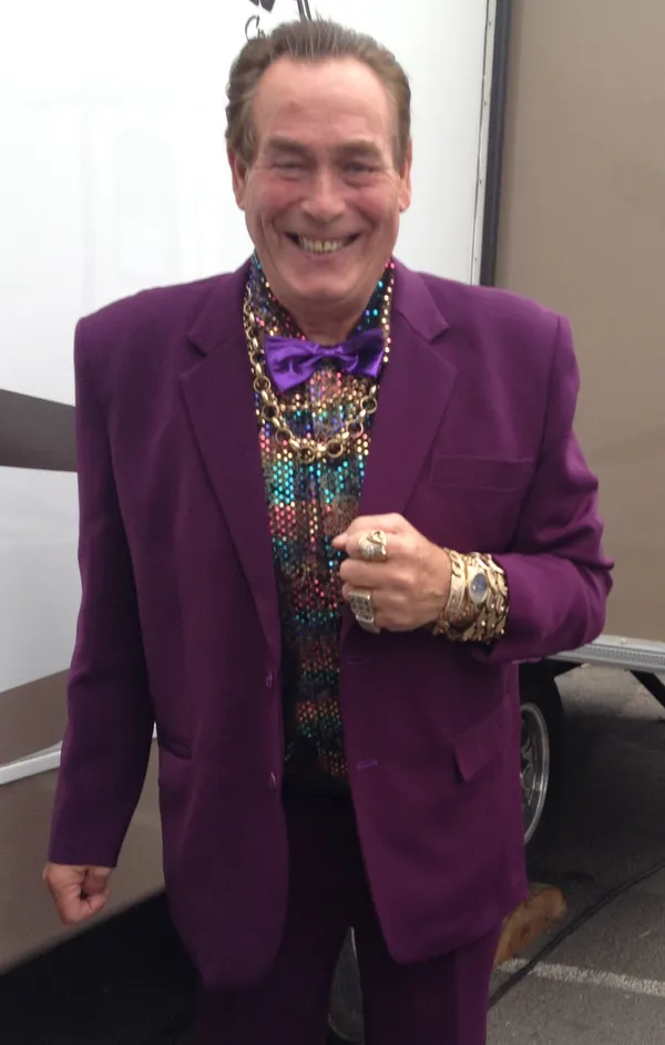英國飛鏢選手 Bobby George 穿紫色西裝、亮片襯衫、戴金鍊微笑，招牌的 King of Bling 打扮