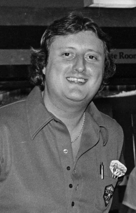 1985年黑白照片中的飛鏢傳奇Eric Bristow笑著面對鏡頭，身穿深色polo衫別著飛鏢徽章