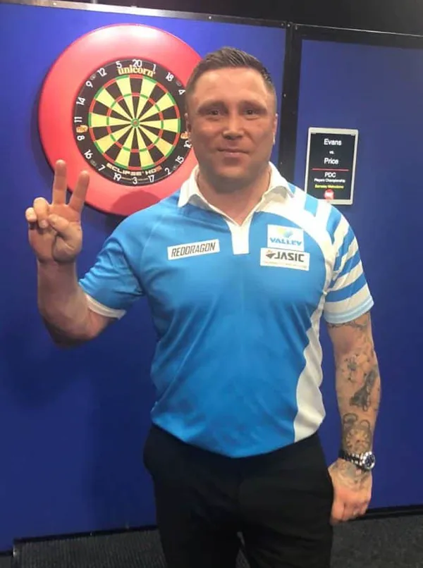 威爾斯飛鏢選手 Gerwyn Price 站在鏢靶前比出勝利手勢,身穿藍白隊服