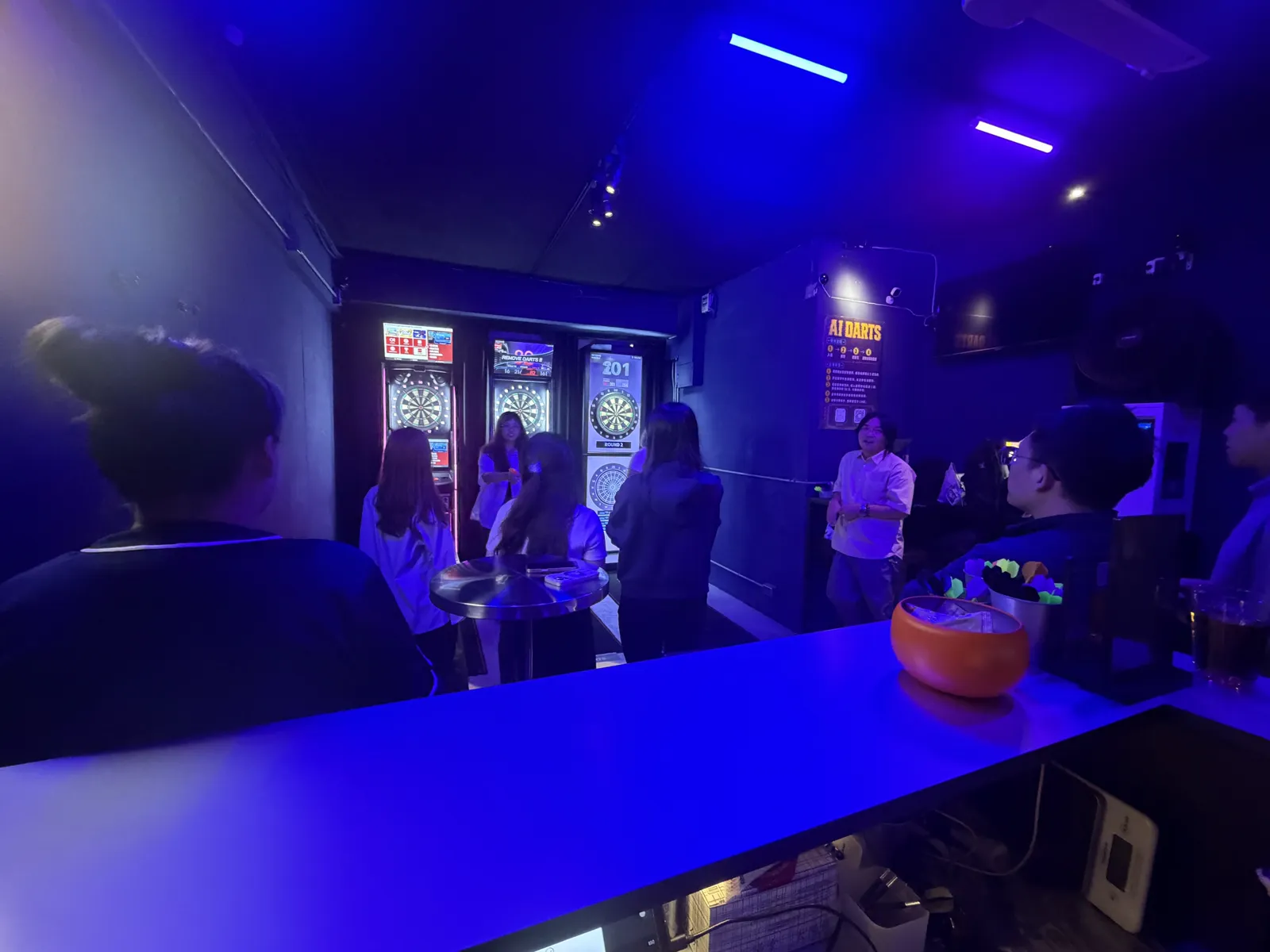 AI DARTS 店內一景，玩家聚在機台前輪流投鏢，牆上有 AI DARTS 說明海報