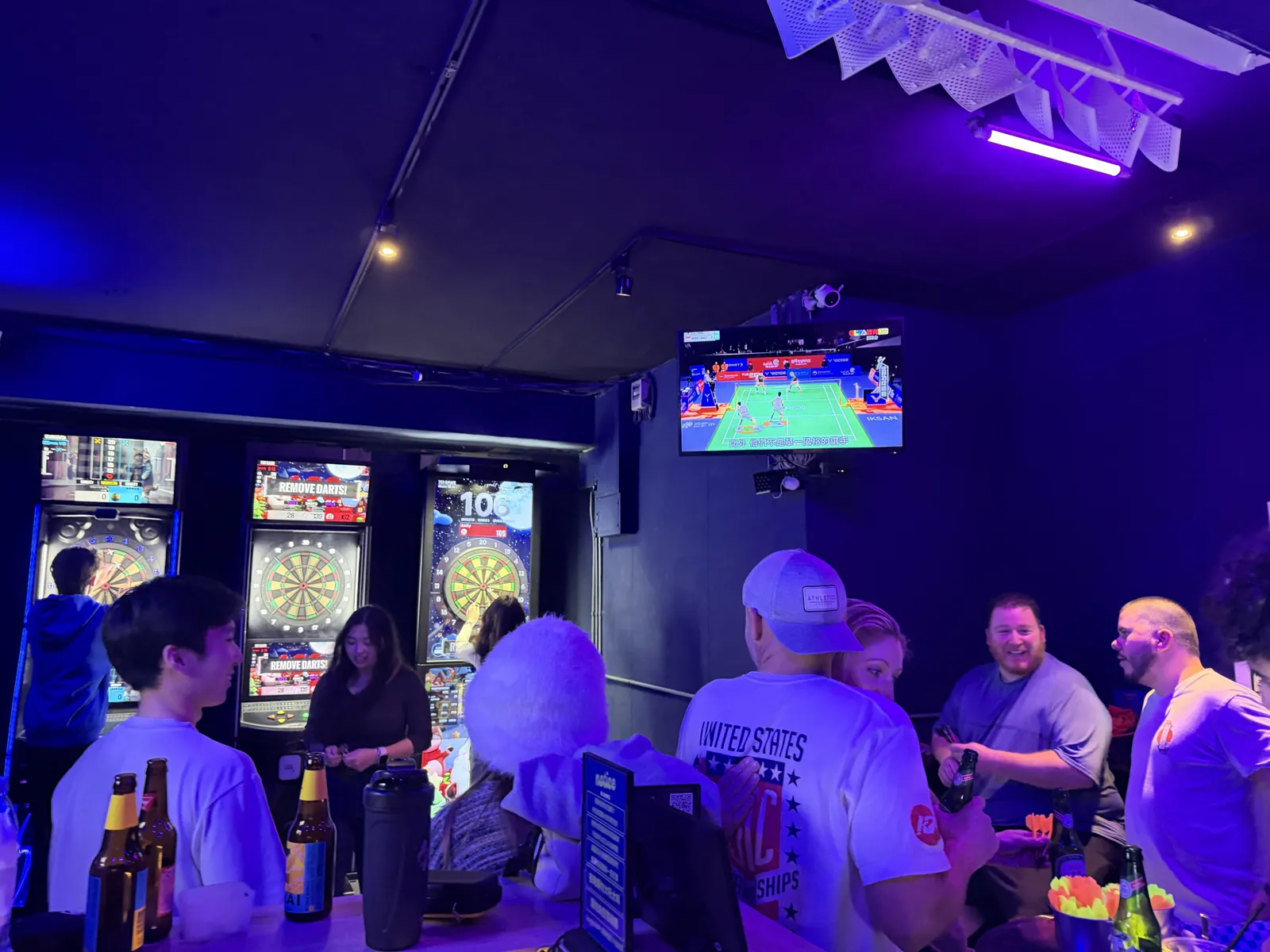 AI DARTS 店內，牆上的電視播著比賽，客人在吧檯邊喝酒聊天，後方機台有人在投鏢